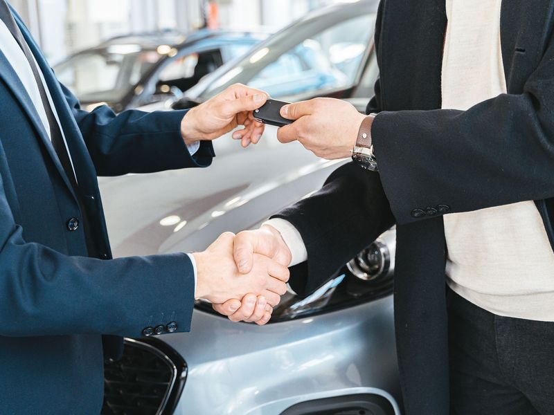 A set of car keys being handed over with a handshake beside a car